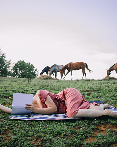 woman laying in a field reading, with horses behind her